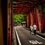 Photo of two people walking across a red iron bridge in Taiwan with greenery and a bus in the background.